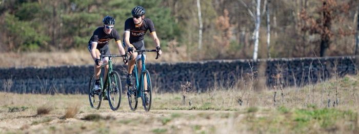 Twee fietsers met 12GO Biking fietskleding op gravelbikes over droog en vlak bospad met bomen en een stenen muur op de achtergrond