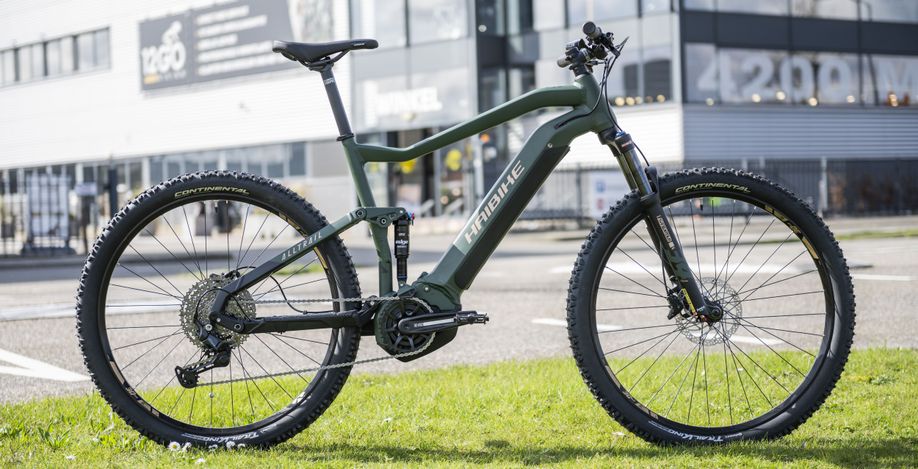 Groene Haibike Alltrail 4 mountainbike op het gras voor een asfaltweg met de winkel van 12GO Biking op de achtergrond