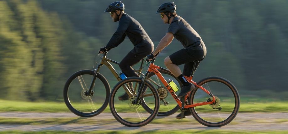 Twee mannelijke fietsers op Cube mountainbikes op gravelpad met wazige bomen op achtergrond en zonlicht vanaf rechts