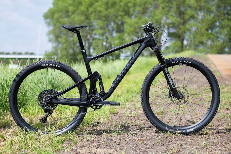 Zwarte Ghost Lector mountainbike vanaf de zijkant, staat op grond en gras naast het fietspad met bomen op achtergrond