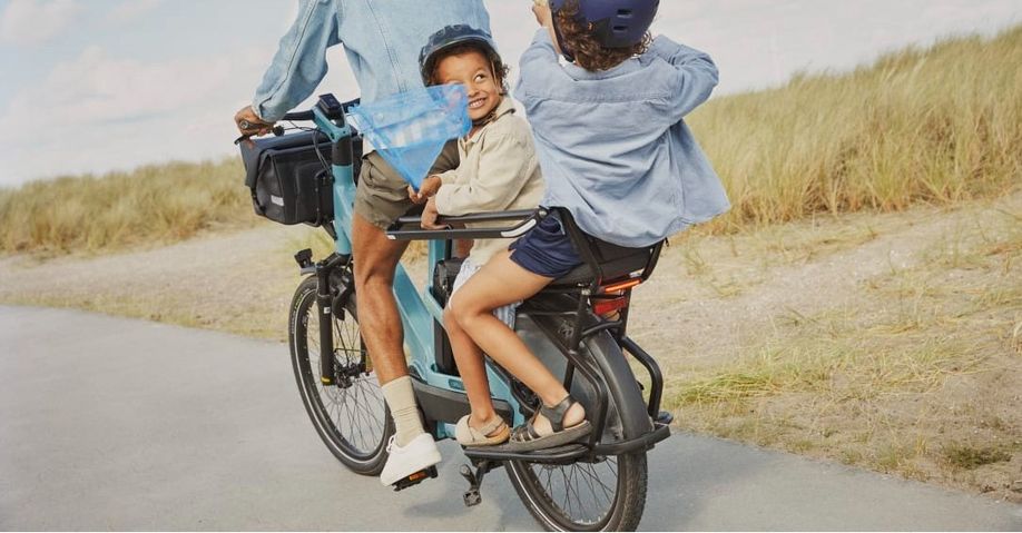 Man met twee jongens achterop blauwe Trek Fetch longtail die met schepnetjes spelen en over een weg langs duingebied fietsen