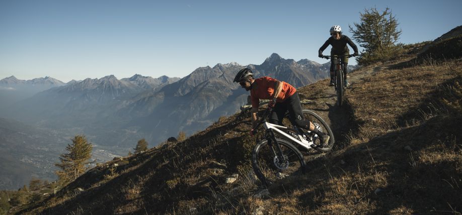 Twee mountainbikers op Scott Genius die van een smal bergpad afdalen met hoge bergen op de achtergrond