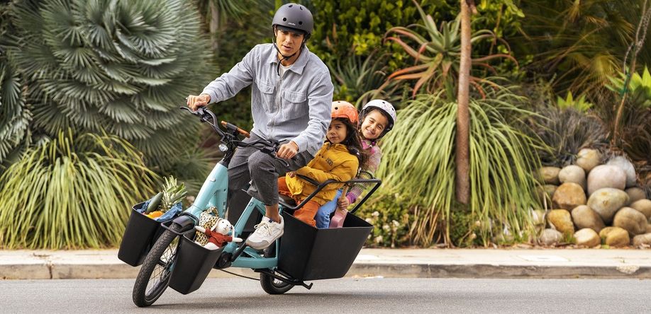 Man met blauwe Trek Fetch longtail hangt iets om een bocht te maken met twee kinderen die achterin lachen voor bijzondere struiken en planten