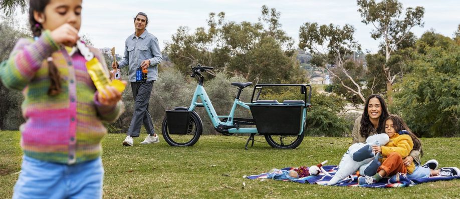 Picknick met vrouw en meisje op kleed, man met spullen uit de blauwe Trek Fetch longtail en meisje met bellenblaas dichtbij de camera