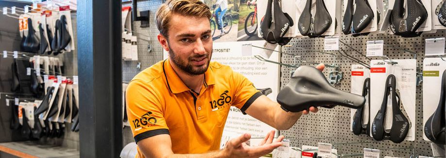 12GO Biking verkoper houdt zadel vast richting de camera voor een rek dat vol met zadels hangt