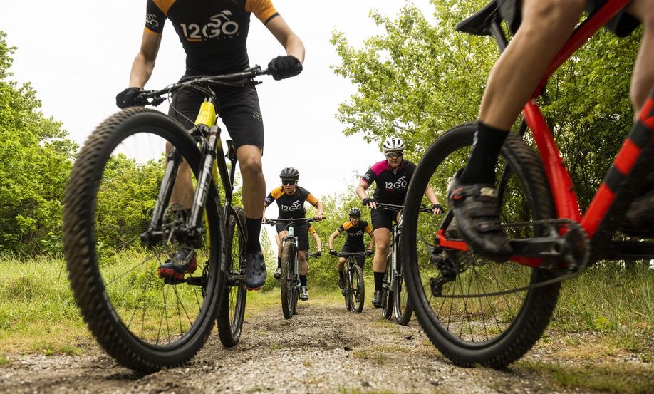 Groep 12GO Biking mountainbikers die aan beide kanten langs de camera fietsen op een grondpad tussen de bomen door