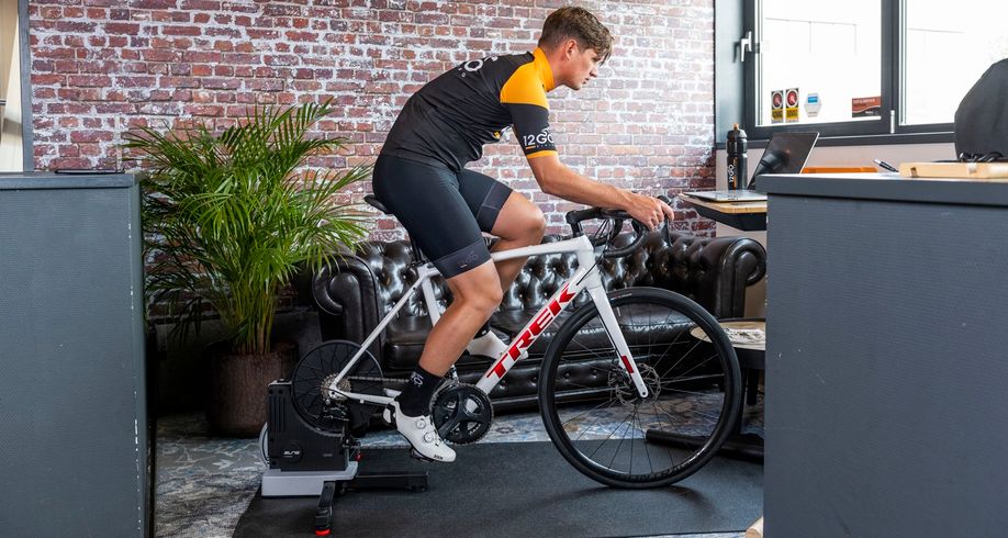 Fietser met 12GO Biking fietskleding op een Elite fietstrainer met witte Trek fiets binnen voor een laptop in werkomgeving