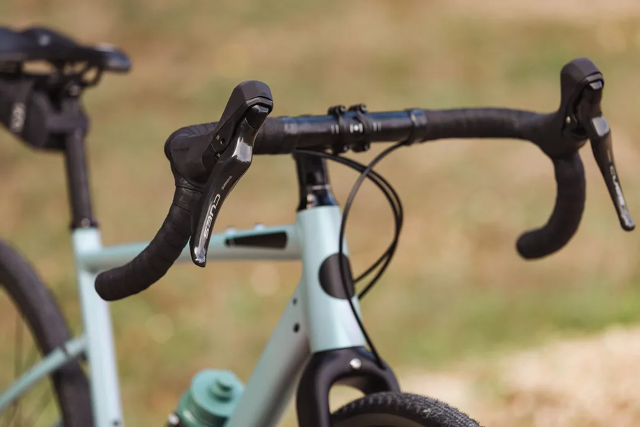 Lichtblauwe gravelbike met bidon en zadeltas heeft Shimano Cues shifters en remmen voor een wazige groen/bruine achtergrond