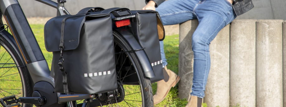 E-bike met dubbele zwarte fietstas achterop en persoon erachter op een betonnen muurtje