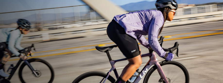 Twee vrouwen staan op de pedalen en komen uit het zadel van twee Liv racefietsen terwijl ze met snelheid op een grote brug rijden