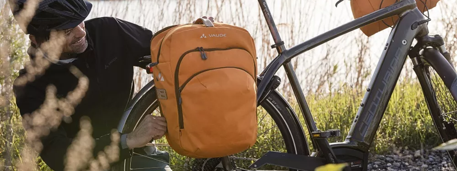 Oranje Vaude enkele fietstas met ritssluiting die door man open gemaakt wordt op een grijze e-bike