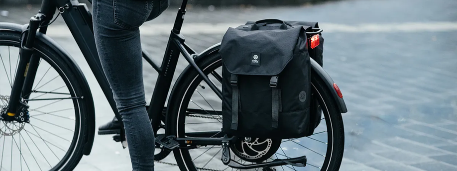 Dubbele zwarte fietstas op een zwarte e-bike met een vrouw en een getegelde straat op de achtergrond