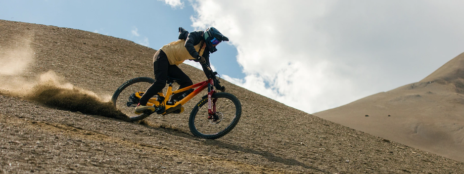 Persoon met full-face helm daalt kale berg af op rood/gele mountainbike waarbij veel stof van de achterkant omhoog komt