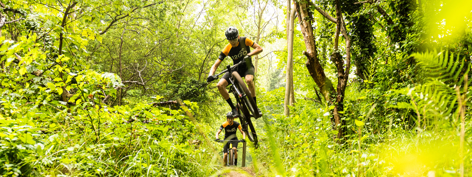 Mountainbikers met 12GO Biking fietsshirt fietsen door het bos richting de camera waarbij de voorste persoon een sprong maakt en in de lucht hangt