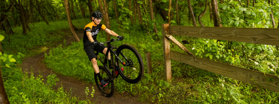 Mountainbiker met 12GO Biking fietsshirt trekt wheelie op bospad met houten hek en veel groene planten op de achtergrond