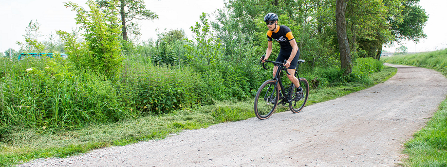 Fietser met 12GO Biking fietskleding rijdt met zwarte gravelbike over gravelpad heen met struiken en gemaaid gras ernaast