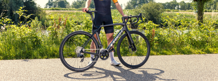Fietser staat met 12GO Biking fietskleding aan en heeft Trek Madone racefiets in zijn hand op asfalt voor gras en bomen