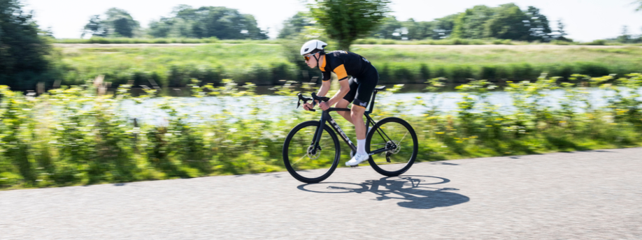 Racefietser met 12GO Biking fietskleding op een zwarte Trek Madone die op asfalt sprint met struiken, een slootje en bomen op de achtergrond