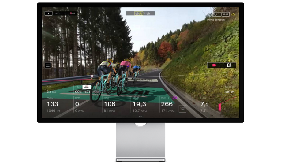 In-game fietsers die een berg afdalen in BKOOL met sponsoren op de weg, bomen links en een wand met gras en planten rechts