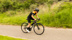 Vrouwelijke fietser in 12GO Biking fietskleding met zwarte racefiets over een fietspad met veel groen gras langs de weg