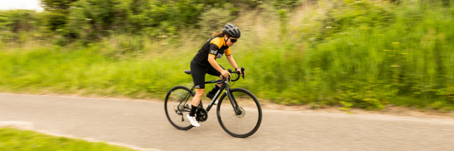 Vrouwelijke fietser in 12GO Biking fietskleding met zwarte racefiets over een fietspad met veel groen gras langs de weg