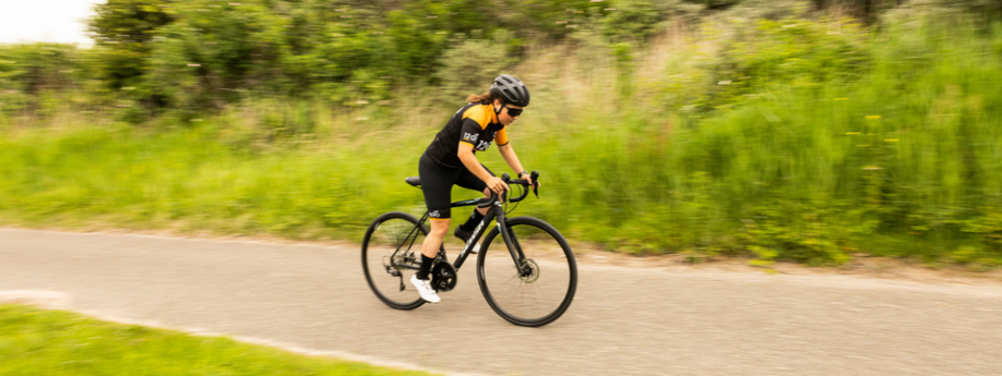Vrouwelijke fietser in 12GO Biking fietskleding met zwarte racefiets over een fietspad met veel groen gras langs de weg