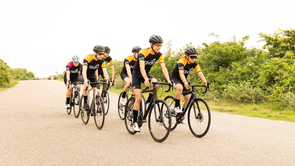 Groep van zes racefietsers met 12GO Biking fietskleding die over een brede weg door de natuurlijk van gras en groene struiken fietsen