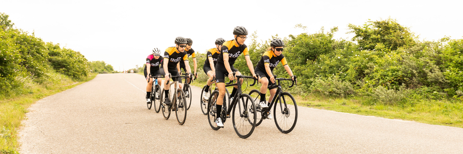 Groep van zes racefietsers met 12GO Biking fietskleding die over een brede weg door de natuurlijk van gras en groene struiken fietsen