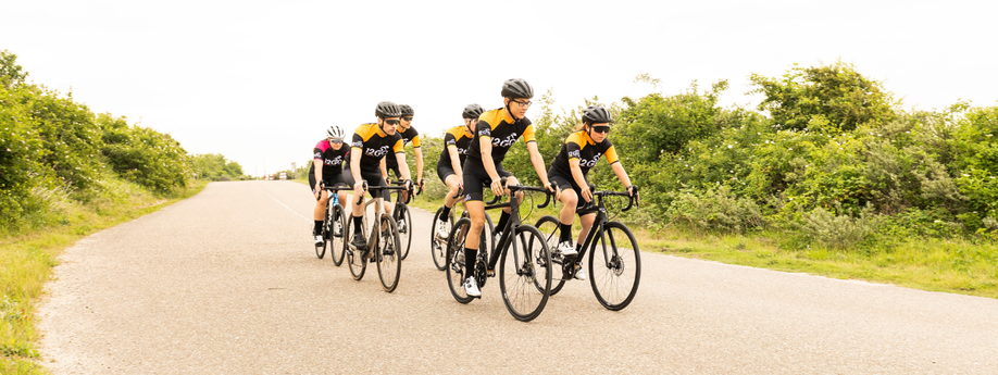 Groep van zes racefietsers met 12GO Biking fietskleding die over een brede weg door de natuurlijk van gras en groene struiken fietsen