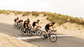 Groep racefietsers met 12GO Biking fietskleding dalen tussen de duinen af over en brede asfaltweg
