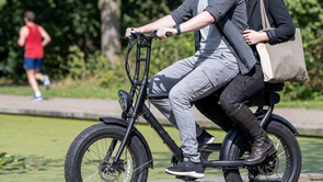 Twee personen samen op een fatbike met verschillende lopers erachter en een fontein met daarachter nog meer mensen op bankjes