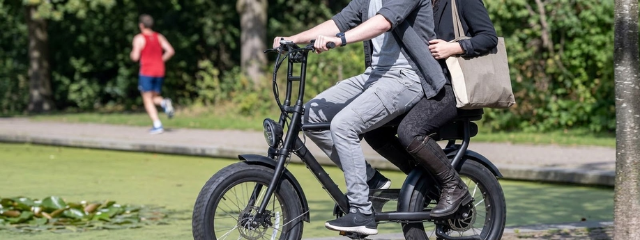 Twee personen samen op een fatbike met verschillende lopers erachter en een fontein met daarachter nog meer mensen op bankjes