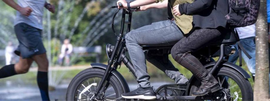 Twee personen samen op een fatbike met verschillende lopers erachter en een fontein met daarachter nog meer mensen op bankjes