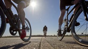 Twee fietsers rijden aan beide kanten dicht de camera langs en een derde fietser rijd over de lijn van het asfalt recht op de camera af met blauwe lucht