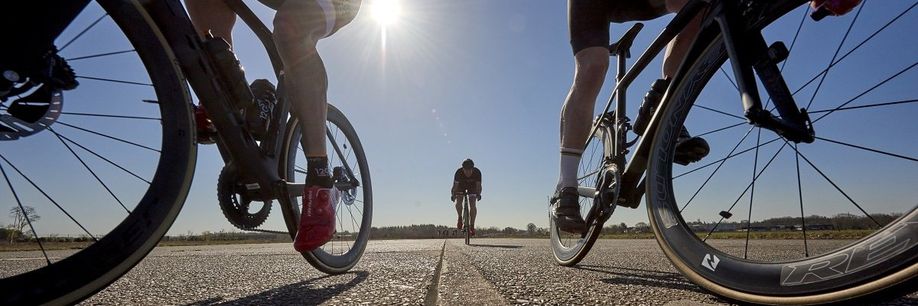 Twee fietsers rijden aan beide kanten dicht de camera langs en een derde fietser rijd over de lijn van het asfalt recht op de camera af met blauwe lucht