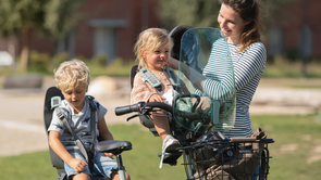 Vrouw met dochter in voorzitje en zoon in achterzitje die de helm van het meisje opzet met een speeltuin en huizen als achtergrond
