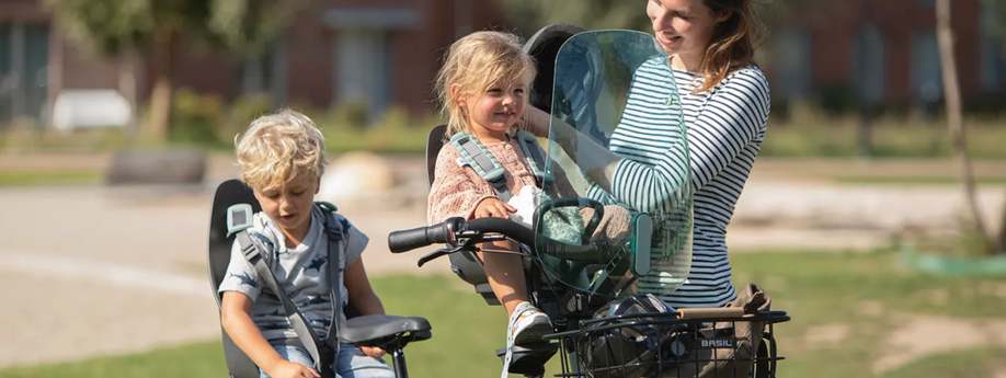 Vrouw met dochter in voorzitje en zoon in achterzitje die de helm van het meisje opzet met een speeltuin en huizen als achtergrond