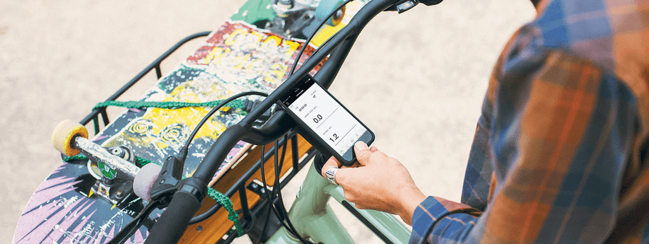 Fietser staat met fiets stil, heeft een skateboard op de mand voor gebonden en heeft telefoon met Cannondale app in haar hand