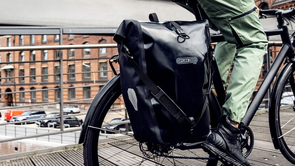 Twee fietsers met beide een gekleurde fietstas voor het rode fietsstoplicht in een stad met unieke huizen op de achtergrond