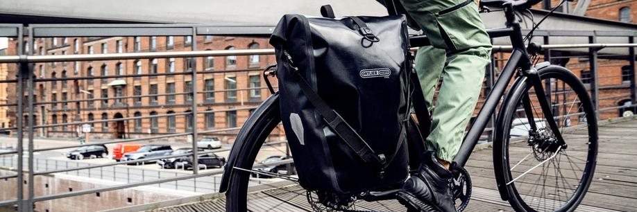 Twee fietsers met beide een gekleurde fietstas voor het rode fietsstoplicht in een stad met unieke huizen op de achtergrond