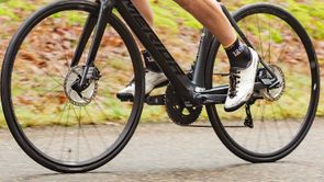 Fietser met 12GO Biking sokken, witte fietsschoenen, zwarte Merida racefiets met schrijfremmen op een asfaltweg voor gras en bladeren