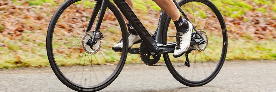Fietser met 12GO Biking sokken, witte fietsschoenen, zwarte Merida racefiets met schrijfremmen op een asfaltweg voor gras en bladeren