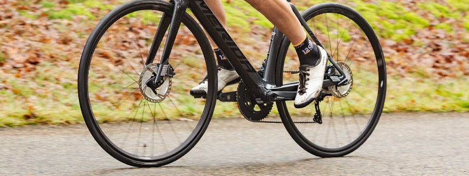 Fietser met 12GO Biking sokken, witte fietsschoenen, zwarte Merida racefiets met schrijfremmen op een asfaltweg voor gras en bladeren