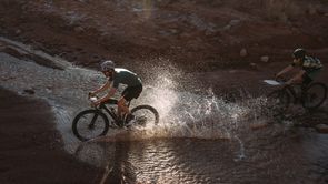 Twee gravelbikers die door het water fietsen om het juiste pad te volgen waarbij de achterste ook nog een kaart voorop de gravelbike heeft zitten
