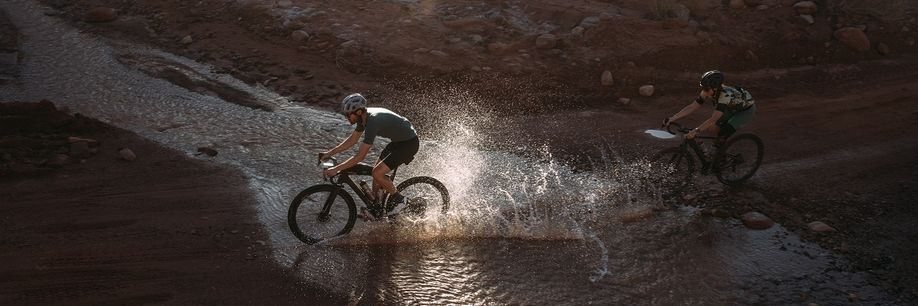 Twee gravelbikers die door het water fietsen om het juiste pad te volgen waarbij de achterste ook nog een kaart voorop de gravelbike heeft zitten