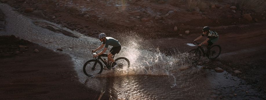 Twee gravelbikers die door het water fietsen om het juiste pad te volgen waarbij de achterste ook nog een kaart voorop de gravelbike heeft zitten