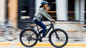 Vrouwelijke fietser met helm op e-bike rijdt snel voorbij huizen met fietsen, bankjes en een hek op de stoep met gele rand