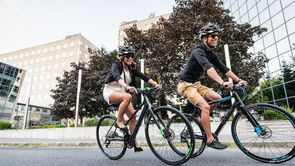 Man en vrouw op zwarte fitness hybride in een stad met bomen en meerdere bedrijfspanden met veel glas op de achtergrond
