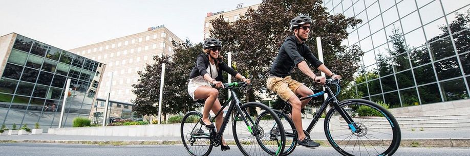 Man en vrouw op zwarte fitness hybride in een stad met bomen en meerdere bedrijfspanden met veel glas op de achtergrond