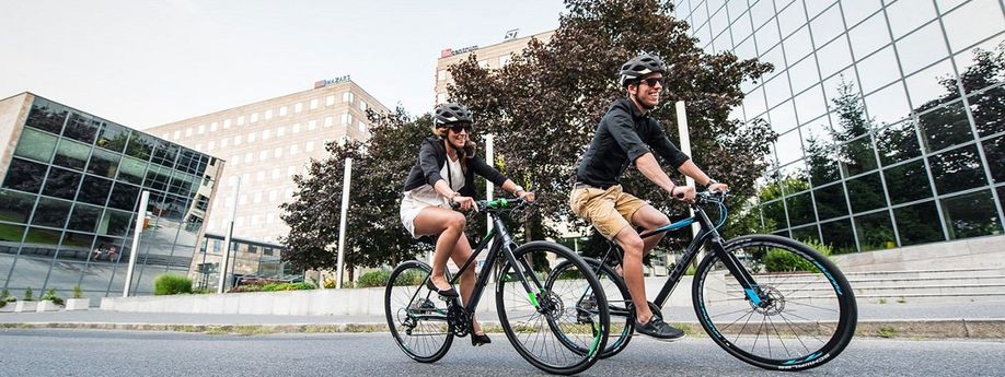 Man en vrouw op zwarte fitness hybride in een stad met bomen en meerdere bedrijfspanden met veel glas op de achtergrond